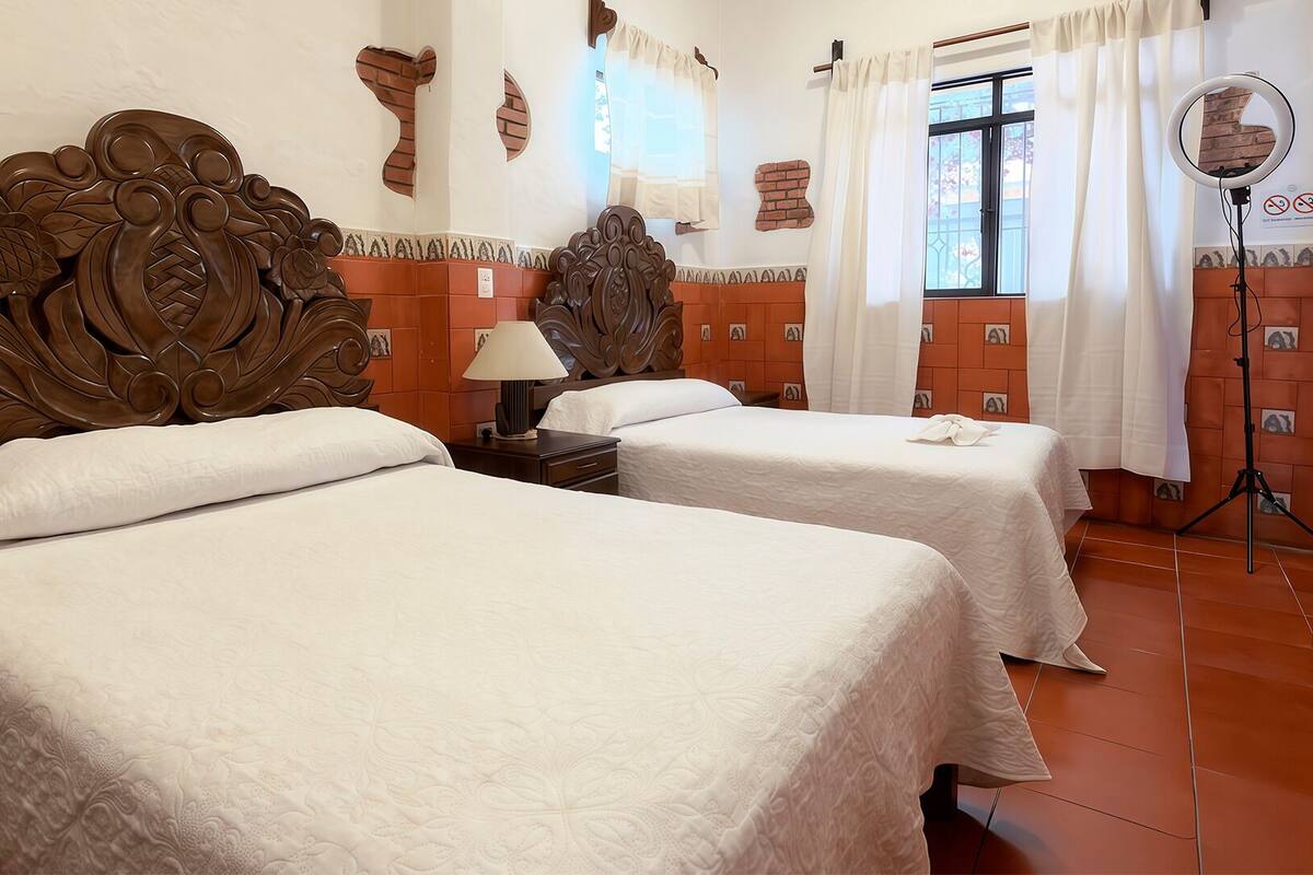 Quarto de hotel com duas camas, cabeceira de madeira, mesa de cabeceira com abajur, espelho e vista para o exterior através de uma janela com cortinas.