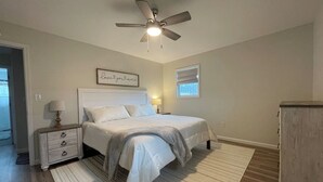 Un dormitorio con cama, mesitas de noche, ventilador de techo, ventana con persianas y una puerta.