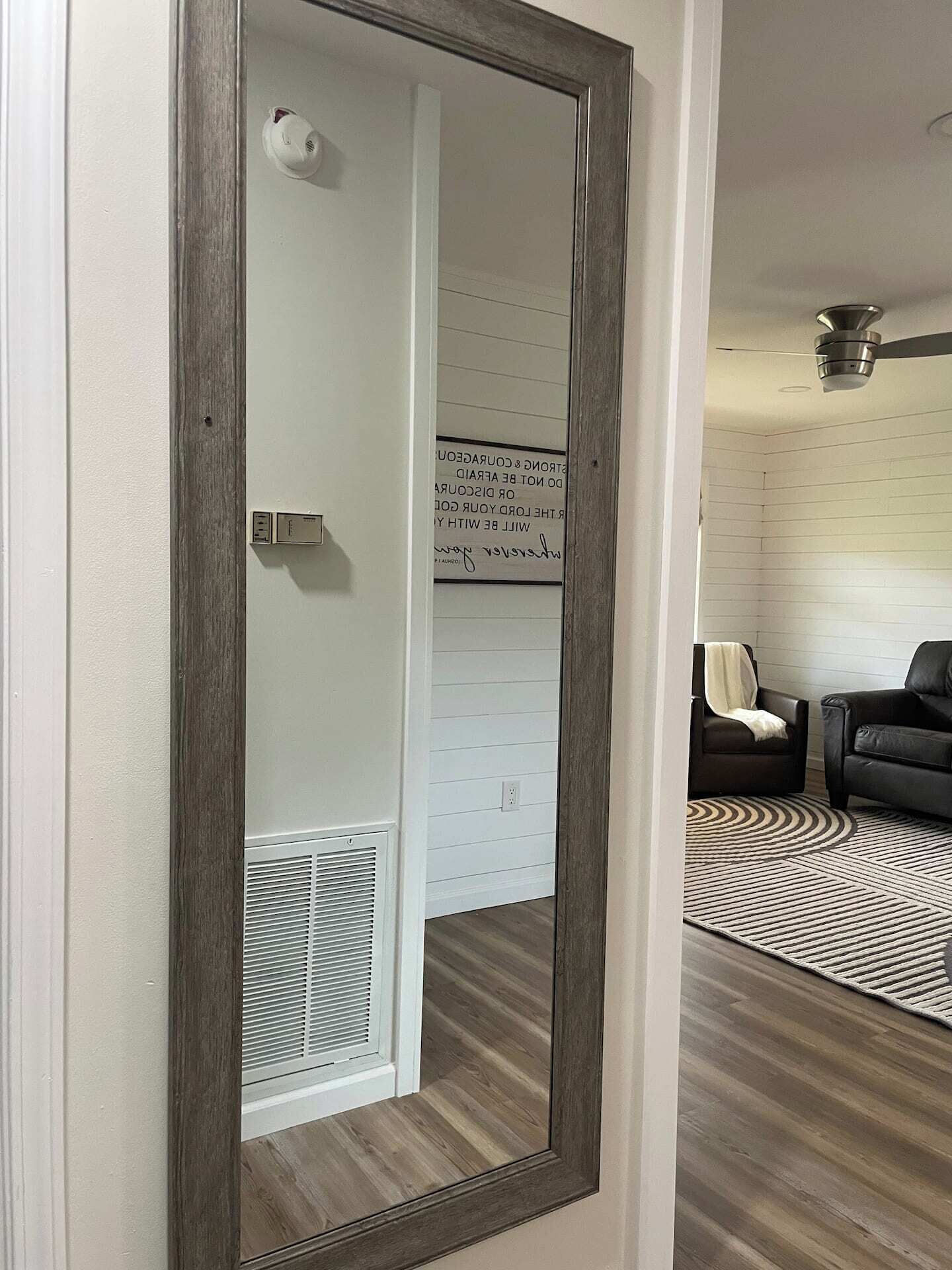 En spegel som reflekterar ett rum med en vit vägg, en svart lädersoffa, en mönstrad matta och en takfläkt.