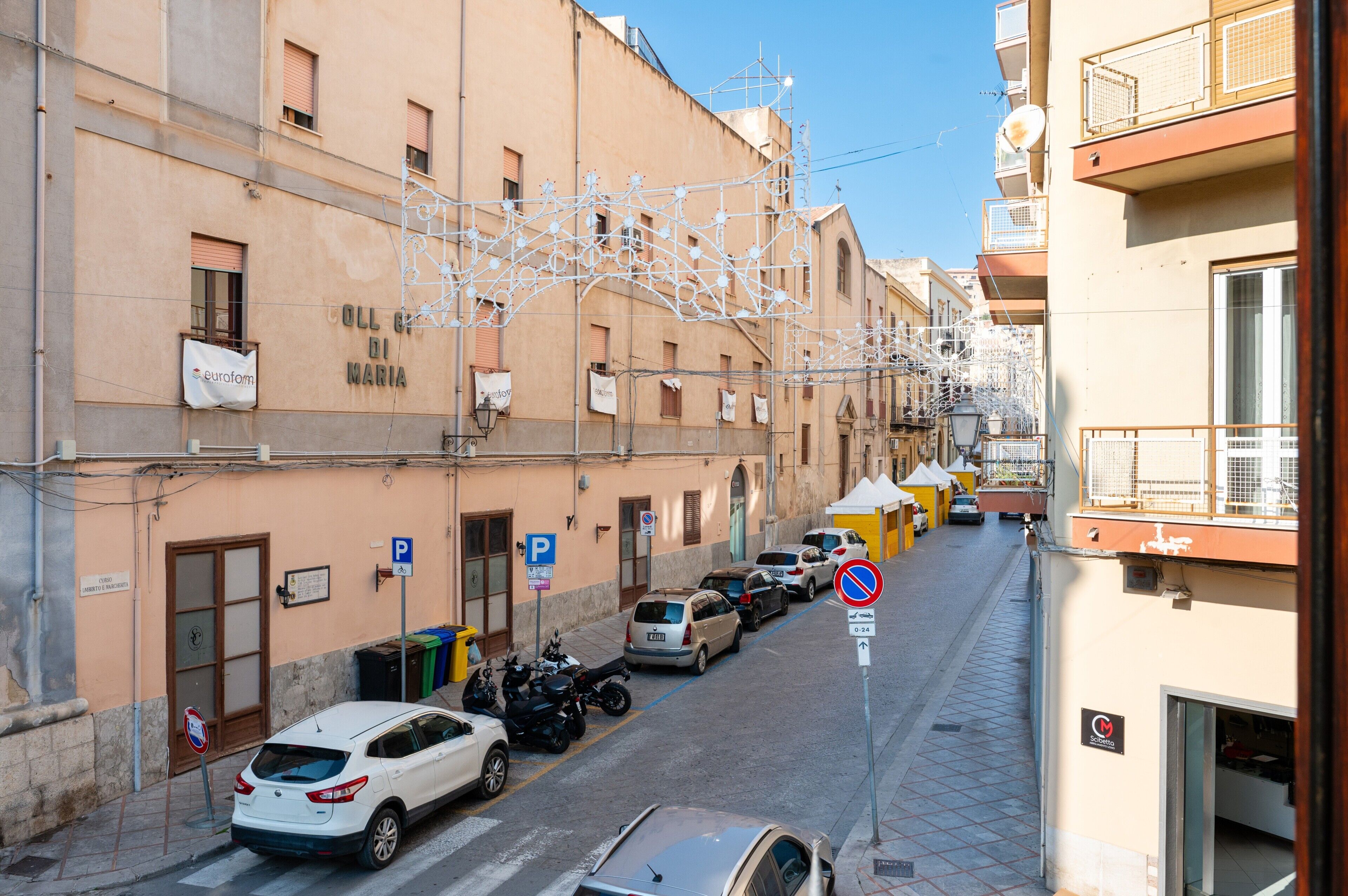 A narrow street lined with parked cars and buildings, including a visible sign for 'COLL. DI MARIA.'