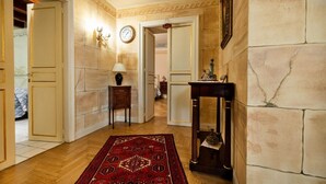 Un pasillo con piso de madera, alfombra roja estampada, un reloj de pared, una mesita con lámpara y una estatua sobre un pedestal.