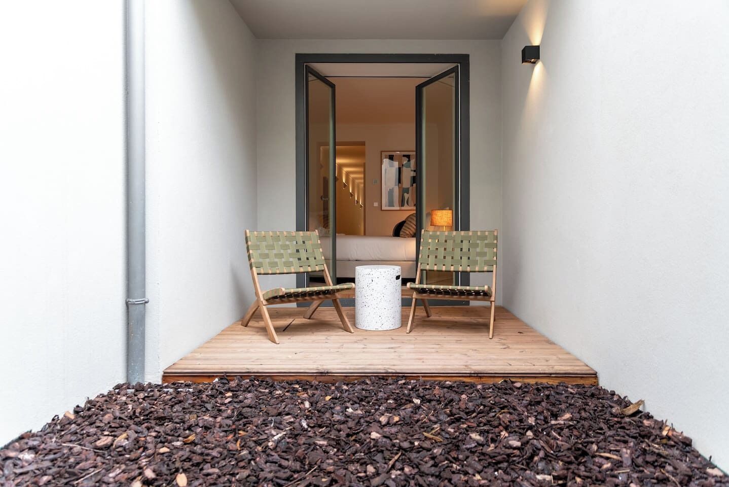 Un petit espace extérieur aménagé avec deux fauteuils en osier et une table blanche, entouré d’une terrasse en bois et d’un tas de paillis.