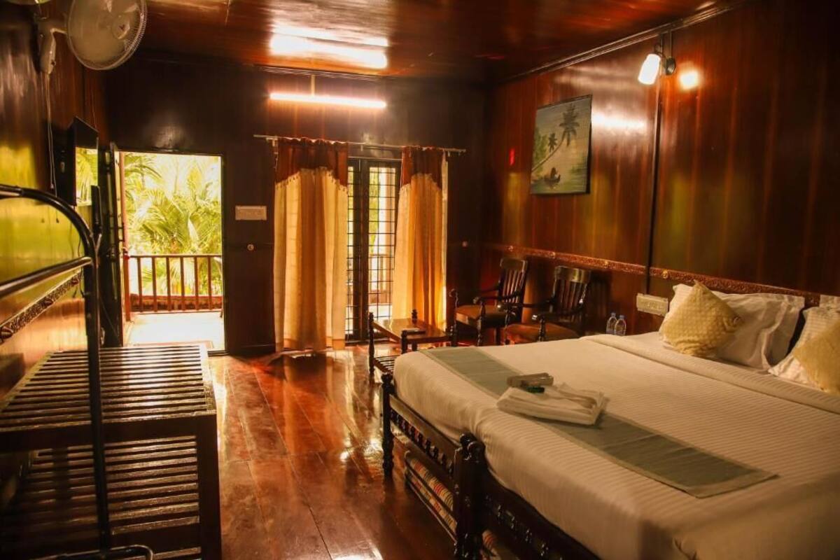 A hotel room with a bed, a desk, and a chair. There is a window with curtains, a door leading outside, and a fan on the wall.