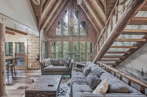 A log cabin interior with a high ceiling, wooden beams, a sofa set, a coffee table, and a view through large windows. - Riverfront Retreat, Sleeps 10・Water Views・Hot Tub (Townsend)