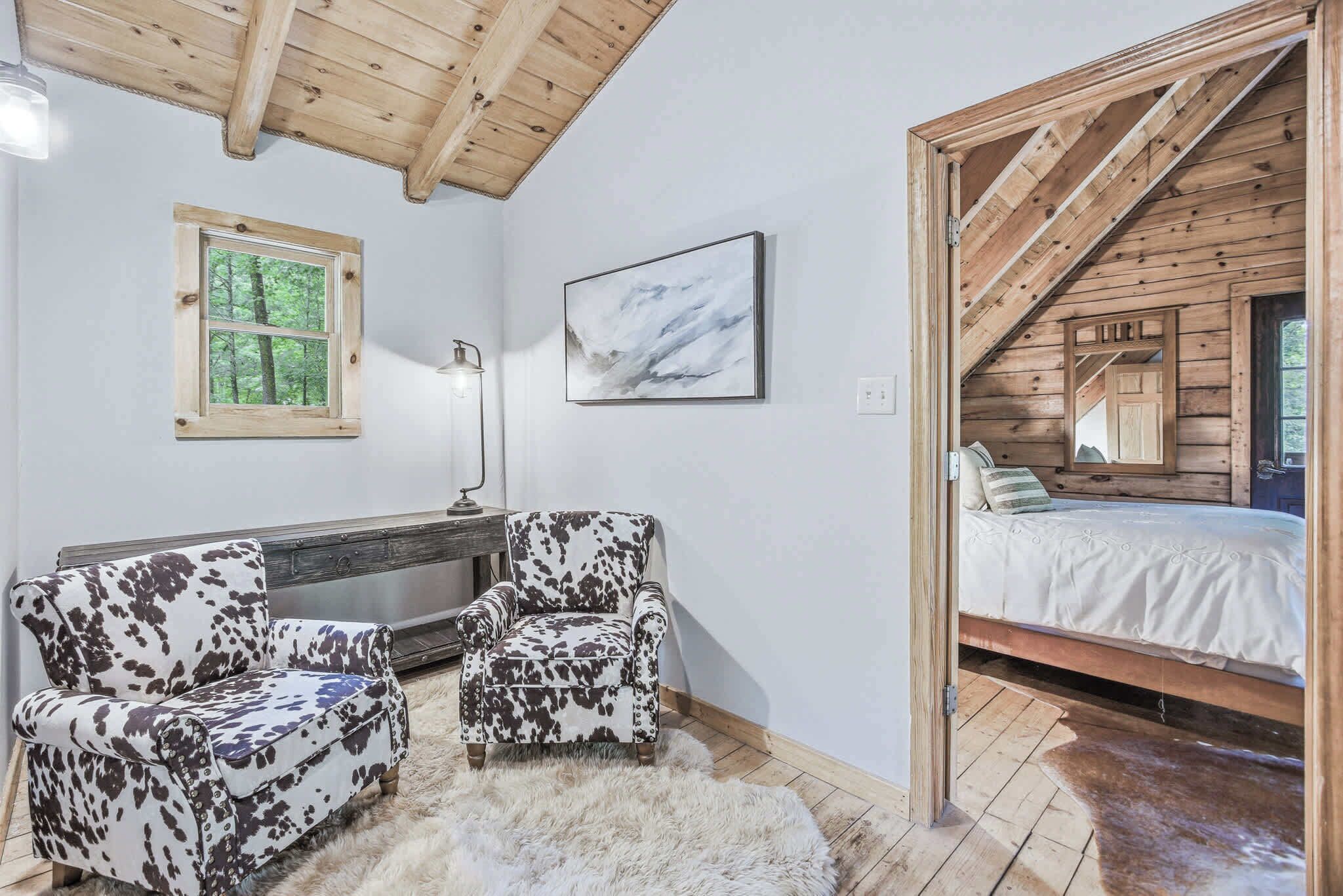 Une pièce confortable avec un lit, deux fauteuils à motifs, une cheminée, une fenêtre donnant sur la forêt et un tableau.