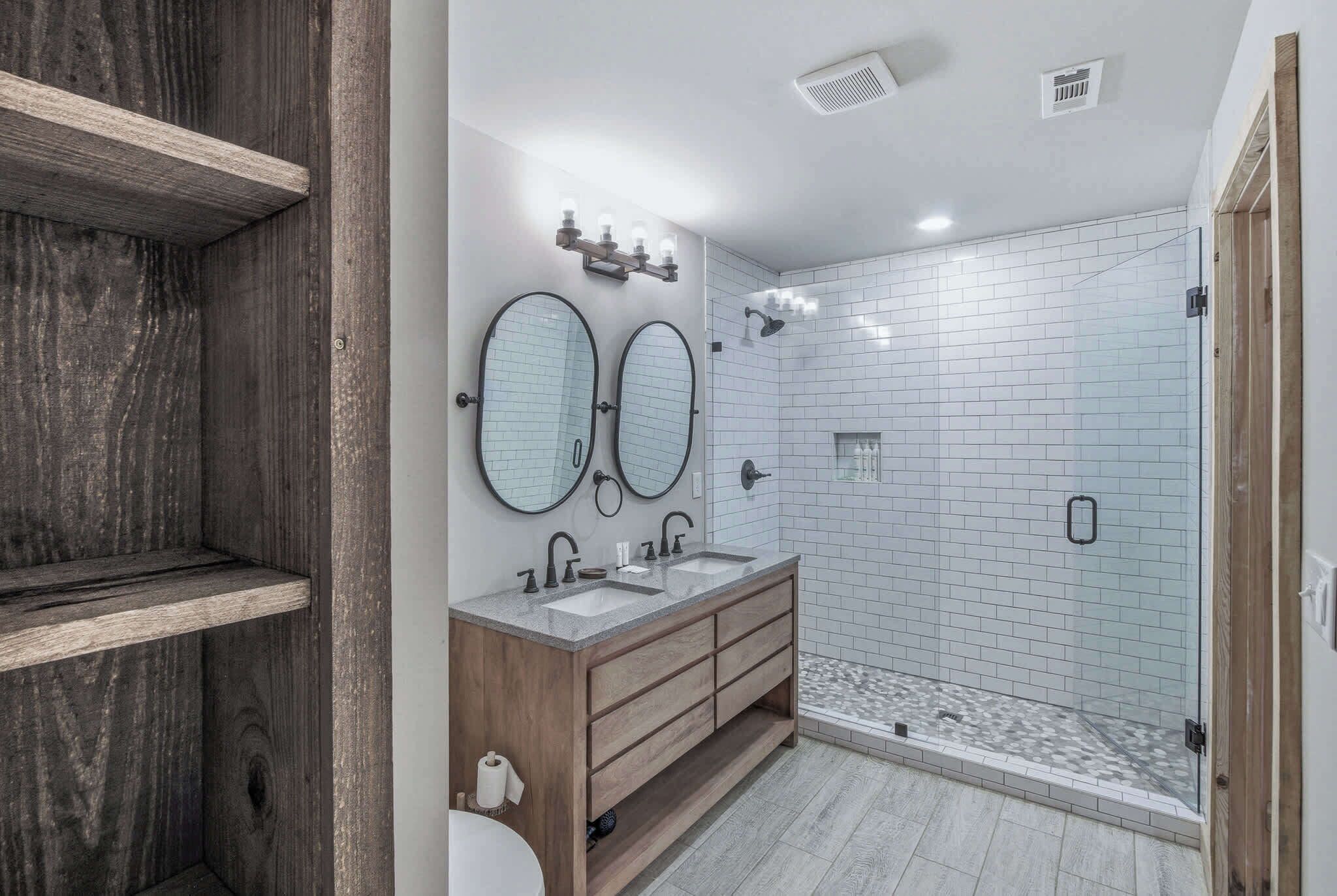 A bathroom with two mirrors, a wooden vanity, a shower with glass doors, and a tiled floor.