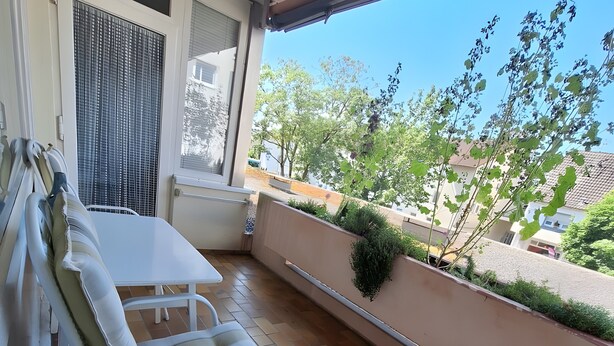 En balkon med et hvidt bord og stole, et vindue med persienner samt udsigt til omkringliggende bygninger og træer.