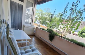 A balcony with a white table and chairs, a window with blinds, and a view of neighboring buildings and trees. - Apartment '58qm' with Balcony and Wi-Fi (Bad Krozingen)