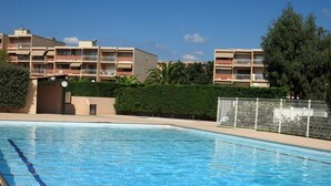 Uma piscina com água azul cristalina, cercada por uma grade e um edifício com várias varandas ao fundo.