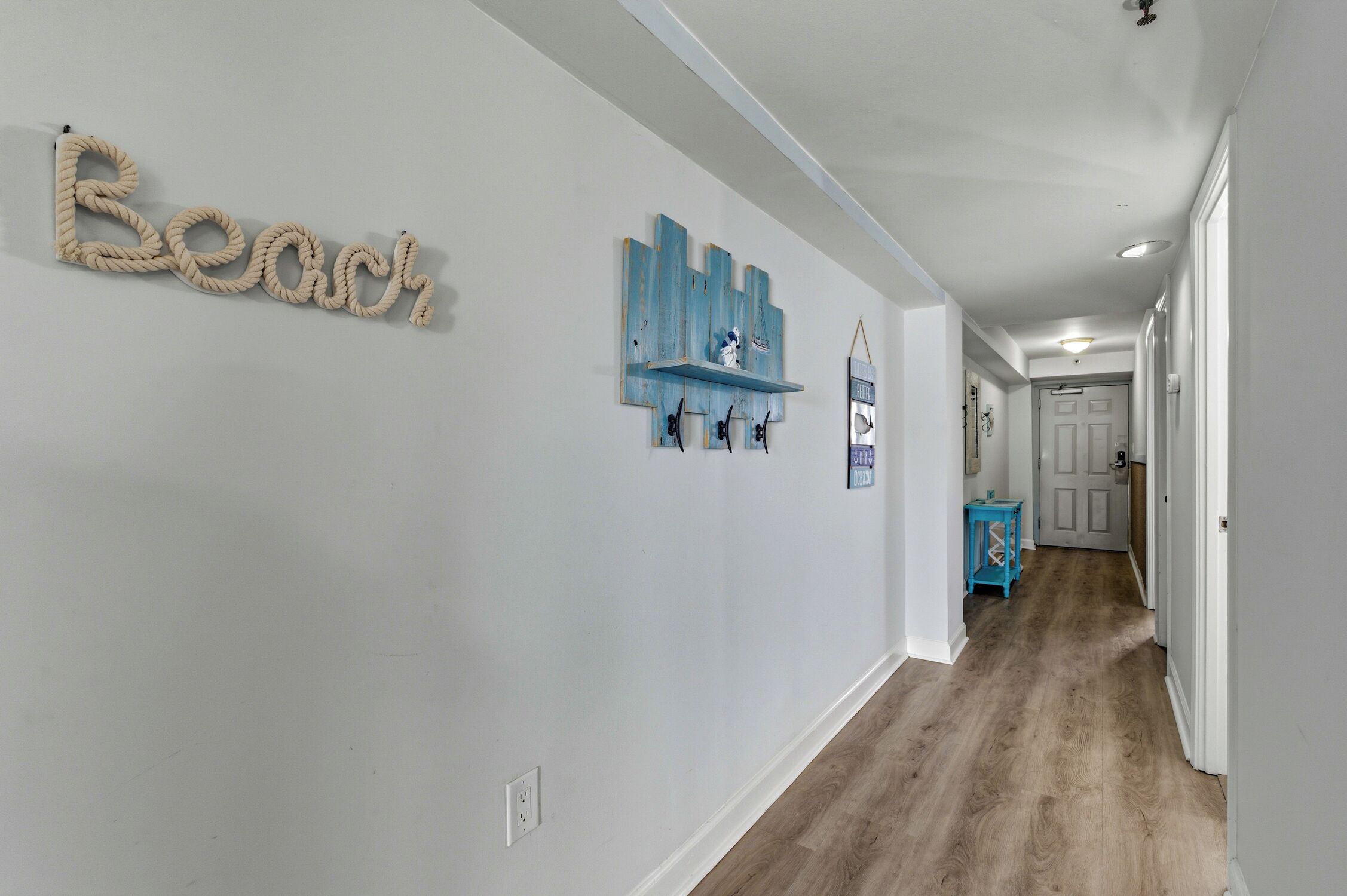 A hallway with a wooden floor, a wall-mounted 'Beach' sign, a blue shelf with decorative items, and a door at the end.