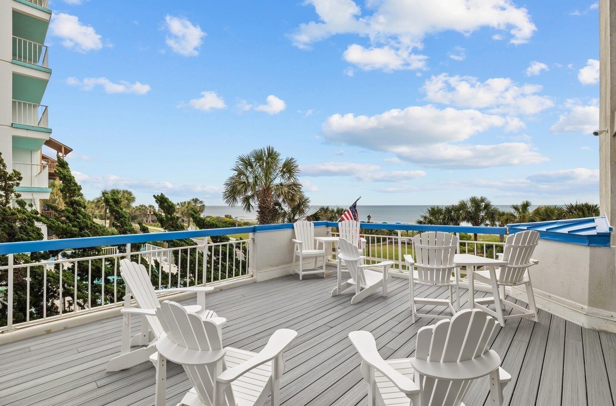 Terraza con sillas Adirondack blancas y una mesa, con vista a una playa y el océano.