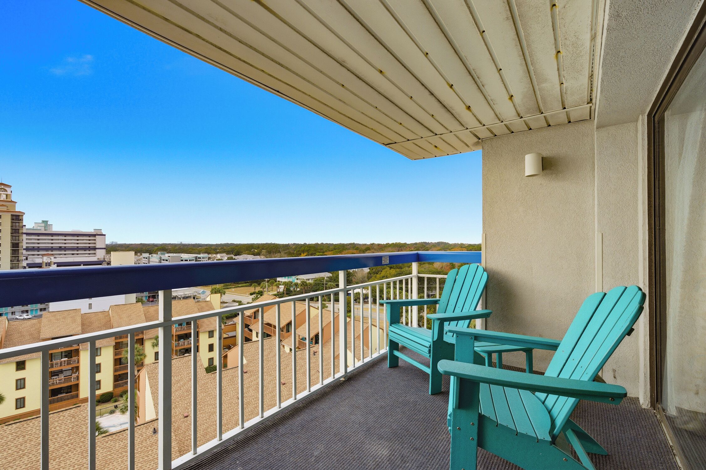 A balcony with two turquoise Adirondack chairs, a view of buildings and a beach, and a clear blue sky.