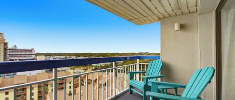 A balcony with two turquoise Adirondack chairs, a view of buildings and a beach, and a clear blue sky.