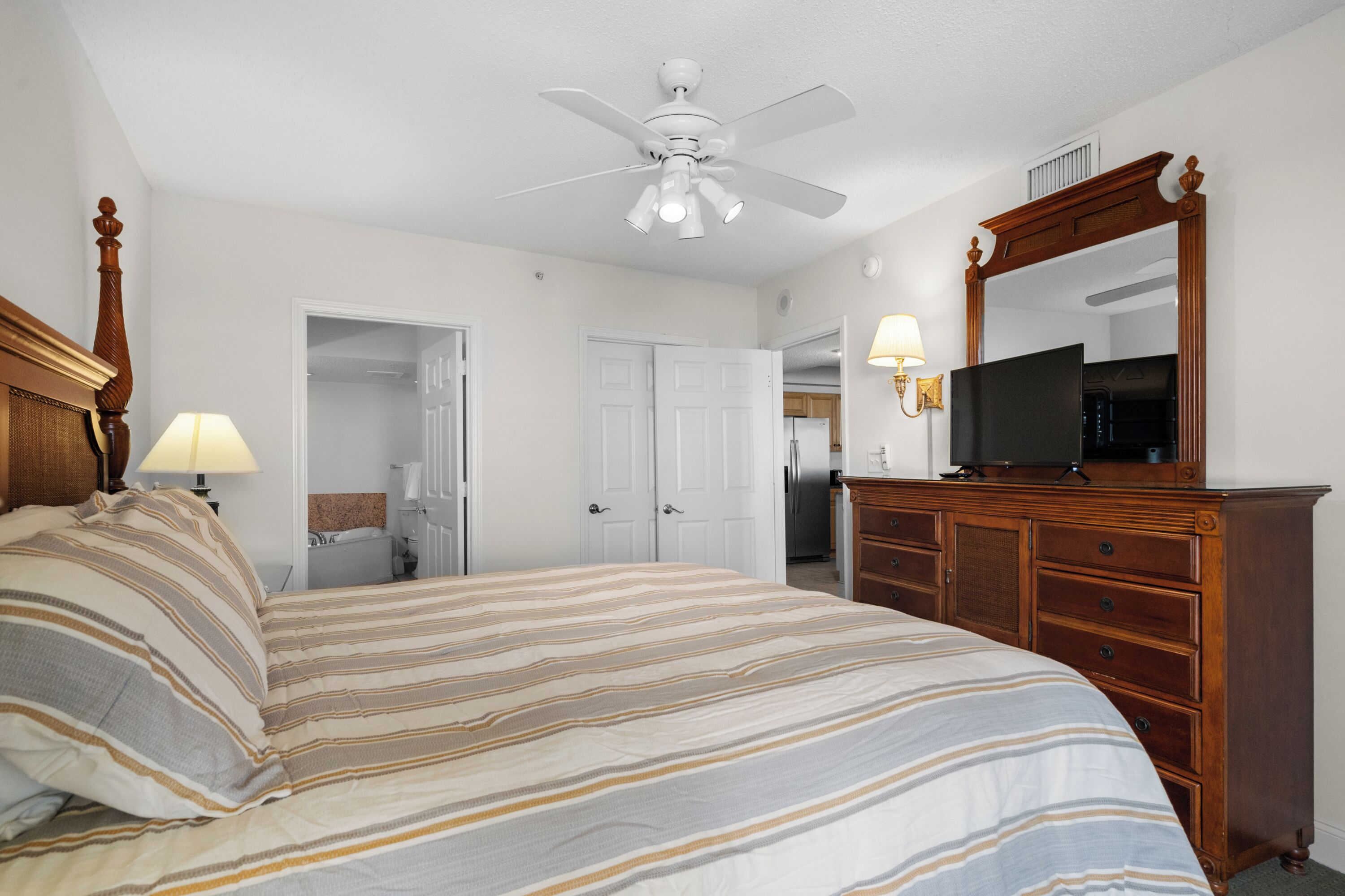 Une chambre à coucher comprenant un lit, une commode en bois, un ventilateur de plafond, un téléviseur posé sur la commode et une porte donnant sur une autre pièce.