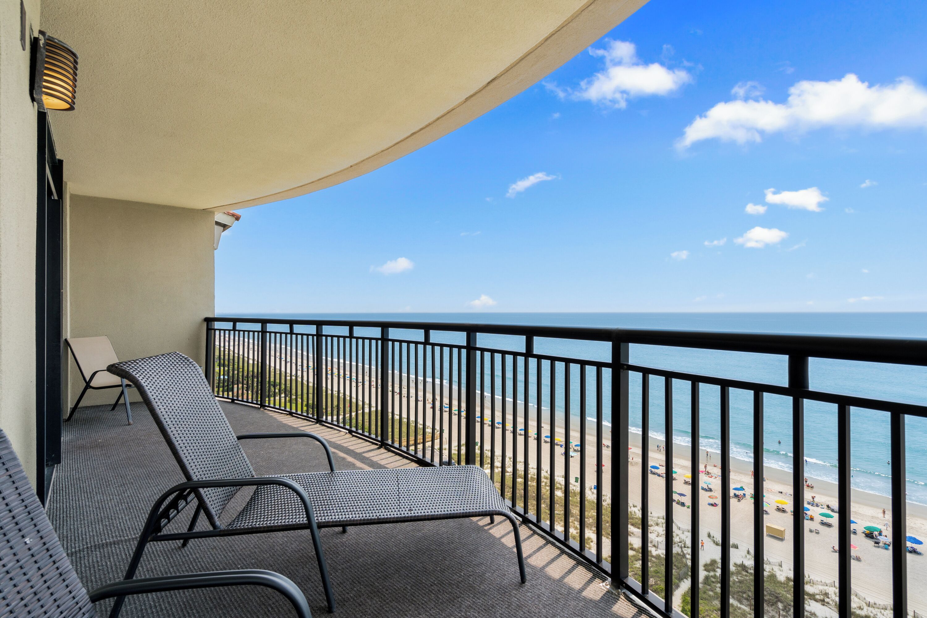 A balcony with two chairs and a table, overlooking a beach with umbrellas and people.