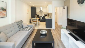 Ein modernes Wohnzimmer mit einer Couch, einem Couchtisch und einem Fernseher. Im Hintergrund ist eine Küche zu sehen.