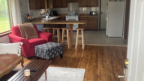 A spacious living room with a wooden floor, a red sofa, a patterned ottoman, and a wooden coffee table. A kitchen area with wooden cabinets, a white refrigerator, and a microwave is visible in the background. - Lovely 2-bedroom apartment in charming Kingsford (Kingsford)