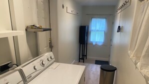 A laundry room with two white washers and dryers, a small trash can, and a window with sheer curtains. - Lovely 2-bedroom apartment in charming Kingsford (Kingsford)