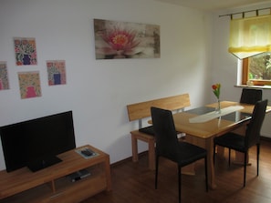 Une pièce comprenant une table à manger en bois, quatre chaises, un banc, un téléviseur posé sur un meuble et des œuvres d’art accrochées au mur.