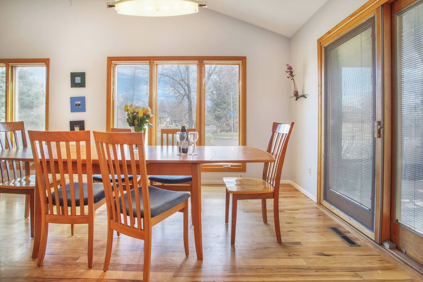 Un comedor con mesa y sillas de madera, ventanas grandes y vistas a árboles.