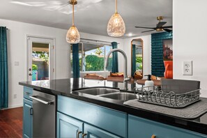 Cocina moderna con encimera negra, pileta de acero inoxidable y alacenas azules. Hay dos lámparas colgantes y un ventilador de techo.