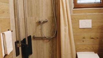 Une salle de bain avec un plancher en bois, une douche équipée d’une barre d’appui, des toilettes et une fenêtre avec des stores.