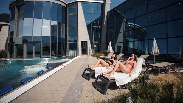 Un edificio moderno con amplios ventanales y un área de piscina con sillones y sombrillas.