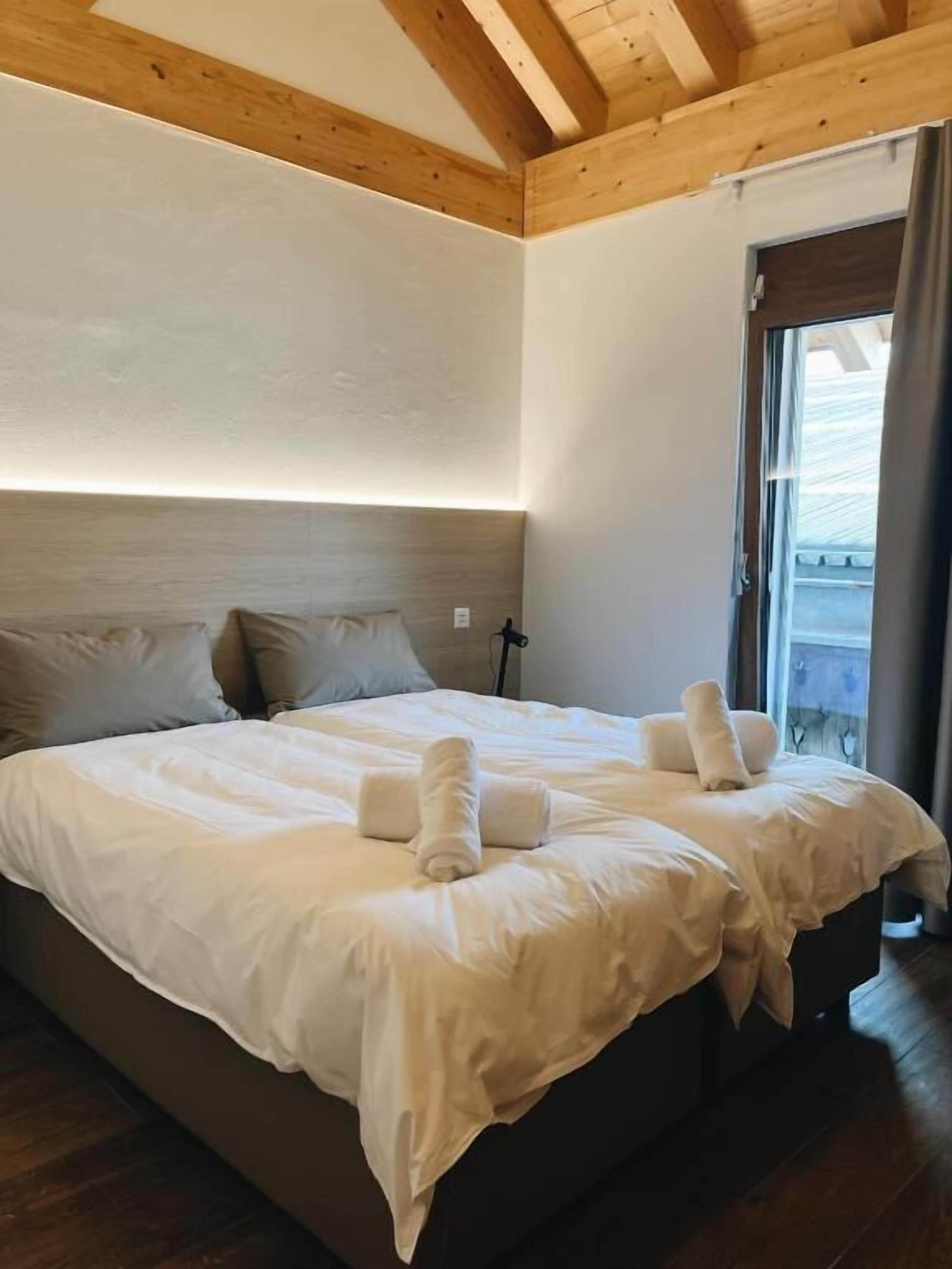 Una cama grande con sábanas blancas y toallas enrolladas, techo de madera y ventana con cortinas.
