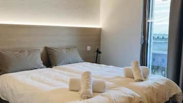 Una cama grande con sábanas blancas y toallas enrolladas, techo de madera y ventana con cortinas.