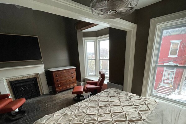 A bedroom with a large bed, a dresser, a chair, a fireplace, and a large window with a view of a red building.