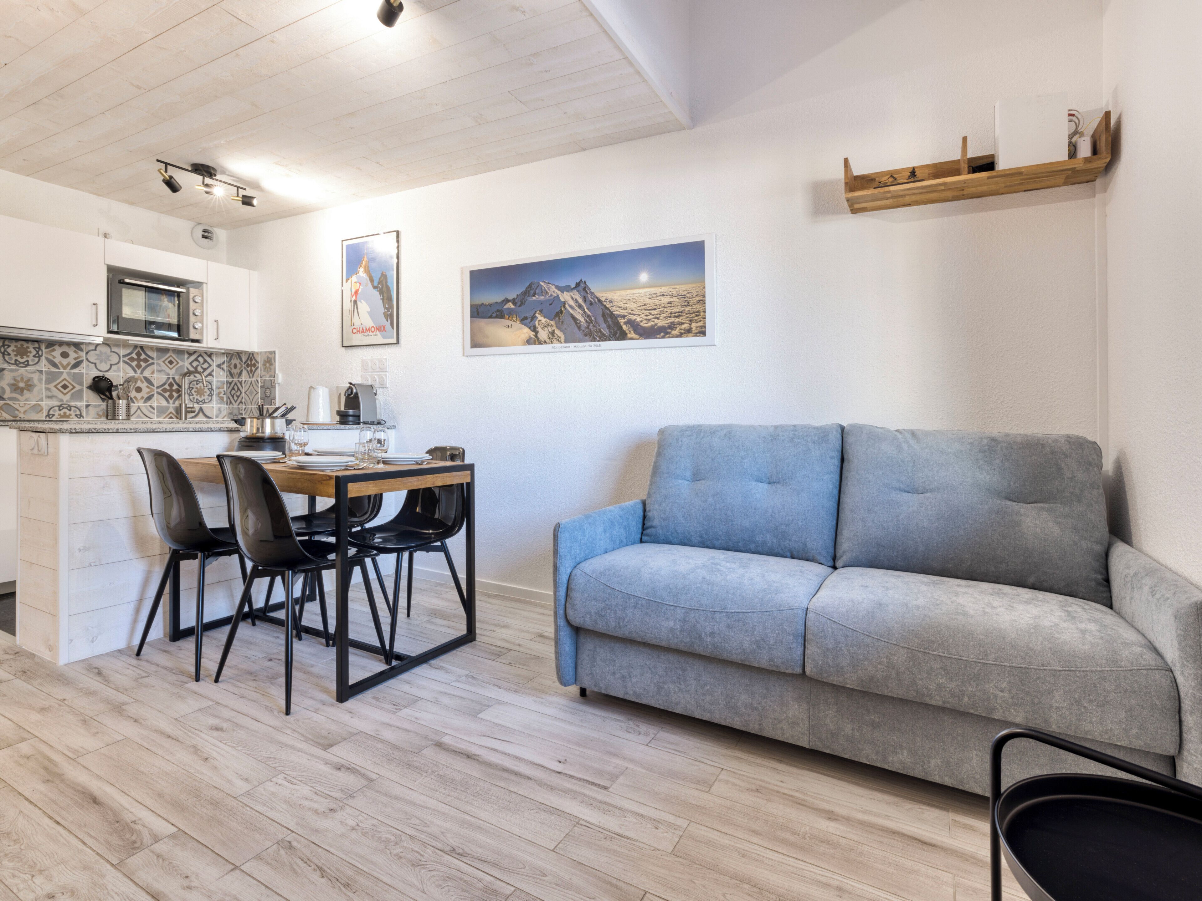 A modern living room with a grey sofa, a wooden dining table with chairs, and a kitchen area with a microwave and tiled backsplash.