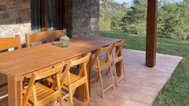 Een terras buiten met een houten tafel en stoelen, stenen muren en uitzicht op bergen.