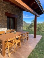 Un patio al aire libre con una mesa y sillas de madera, paredes de piedra y vistas a montañas.