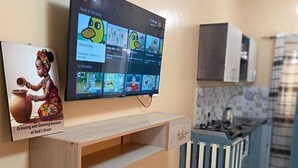 A kitchen with a TV mounted on the wall displaying various channels, a wooden shelf below it, a pink basket on the floor, and a tiled floor with a geometric pattern. - Enjoyable bed & breakfast in charming Diani near the beach (DIANI)