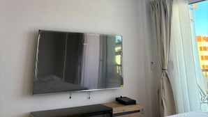 Camera d'albergo con ventaglio a soffitto, TV a schermo piatto fissata al muro, un letto con materasso bianco e una finestra con tende.