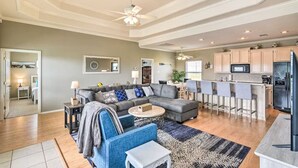 A spacious living room with a gray sectional sofa, a coffee table, and a blue armchair. A kitchen with a bar area is visible in the background. - Family Haven with Fireplace, Garage hangout, and pet friendly (Bolivar Peninsula)