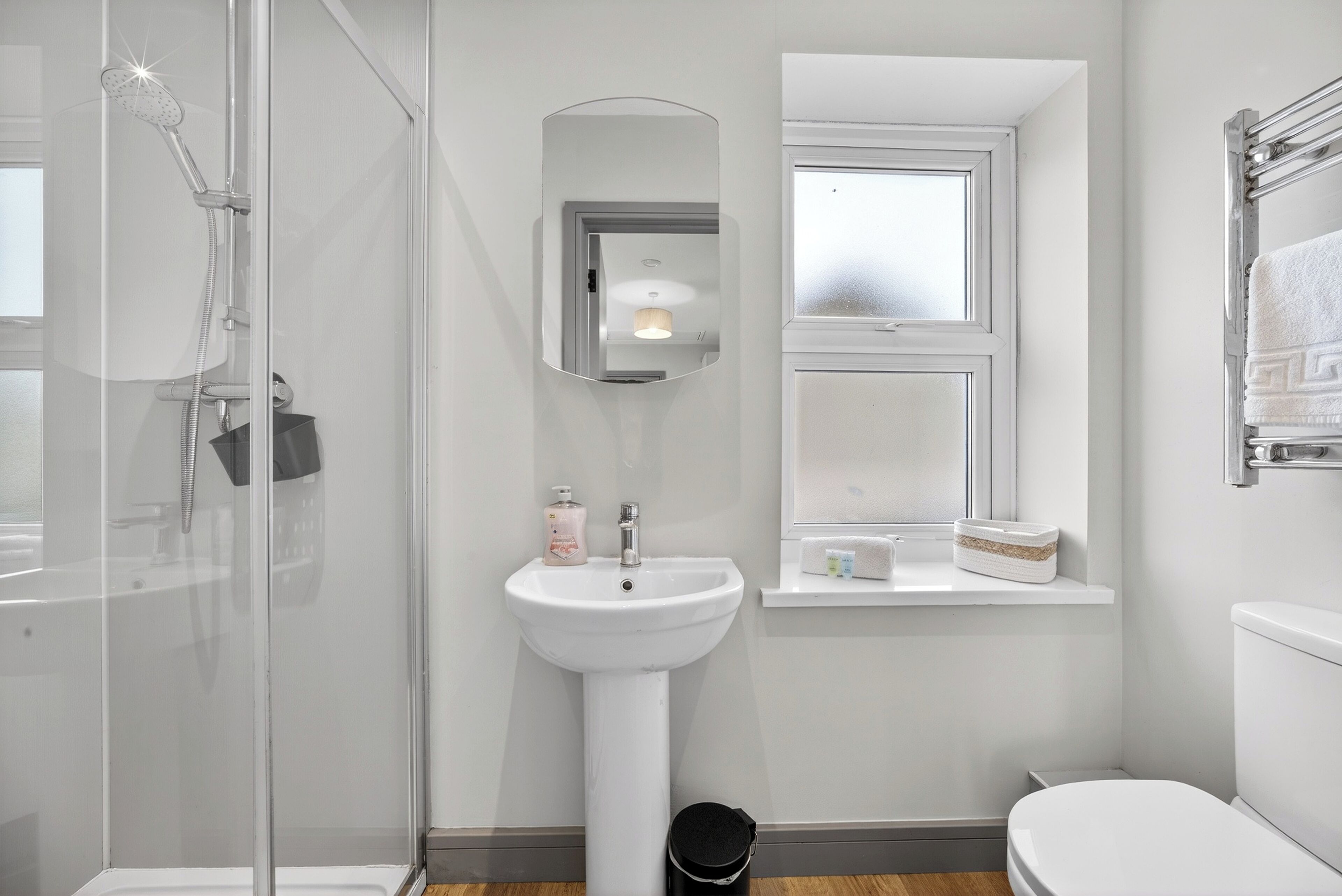 Un baño moderno con ducha de vidrio, lavamanos pedestal, espejo y ventana.
