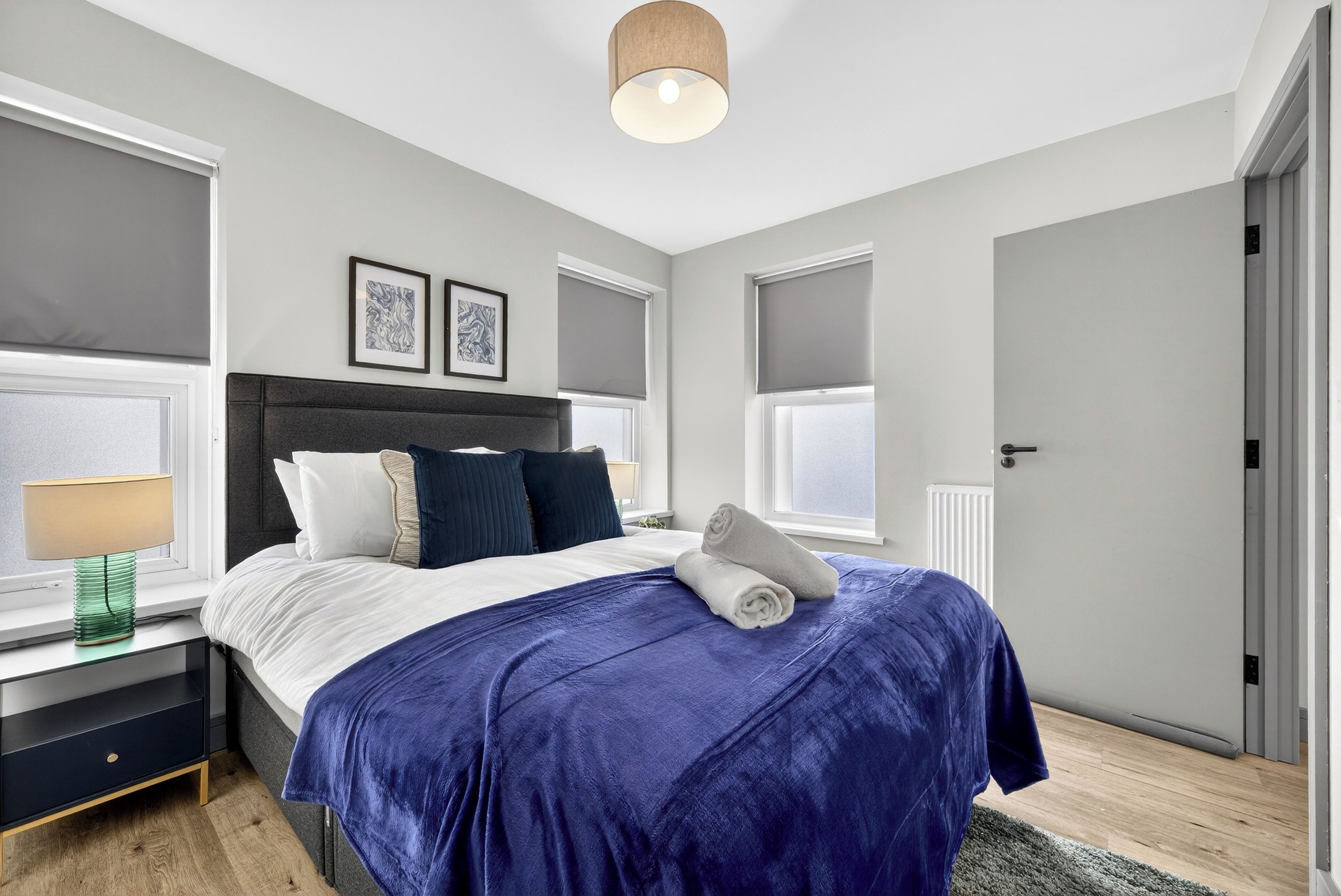 A modern bedroom with a large bed, a bedside table with a lamp, and a window with blinds.