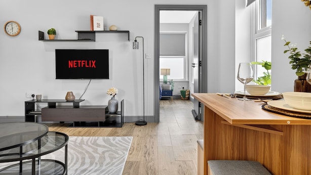A modern living room with a flat-screen TV mounted on the wall, a wooden dining table, and a kitchen area with a wooden countertop and a vase. - Cosy Flat in City Centre (Cardiff)