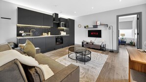 A modern kitchen with dark cabinetry, a microwave, and a built-in oven. There is a round glass coffee table in the living area, a sofa, and a flat-screen TV mounted on the wall. - Cosy Flat in City Centre (Cardiff)