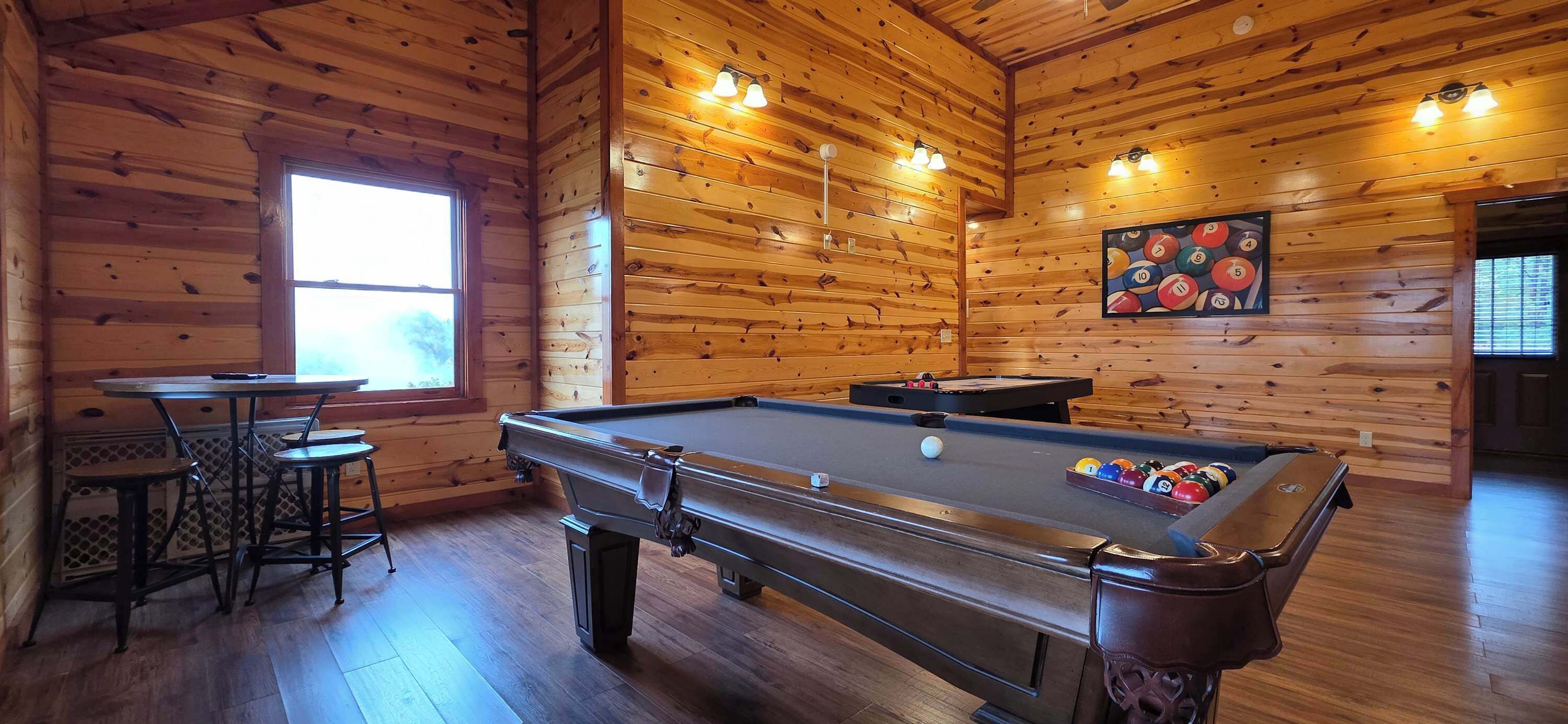 Une table de billard dans une pièce en bois, avec une fenêtre et un tableau représentant des billes de billard.