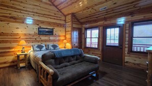 Acogedora habitación de cabaña con paredes de madera, una cama con manta azul, un sillón de cuero, dos lámparas de noche y un ventanal con persianas.