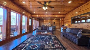 A log cabin interior with a wooden ceiling, large windows, a ceiling fan, a patterned rug, and a leather sofa. - Luxe Cabin, Sleeps 14・Indoor Pool・Hot Tub・Games (Gatlinburg)