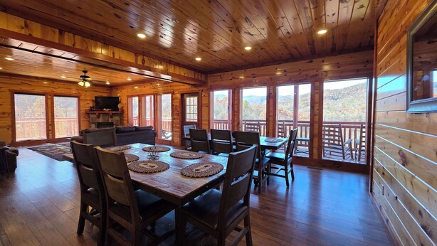 Interior de una cabaña de madera con comedor, chimenea y grandes ventanales con vistas a las montañas.