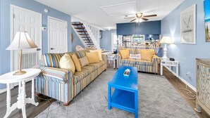 A living room with a striped sofa, a blue coffee table, and a staircase leading to a kitchen. - Laguna Beach Breeze (Panama City Beach)
