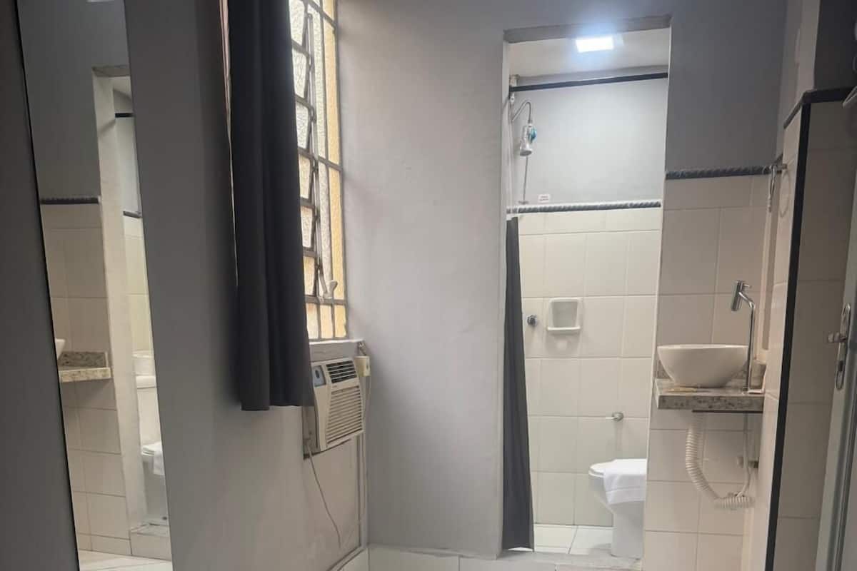 A small, clean bathroom with a shower, toilet, and sink. There is a bed with a rolled towel on it.