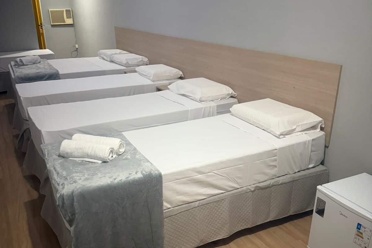 A hotel room with four single beds, each with a grey blanket and white pillows. There is a small white refrigerator on the right side of the room.