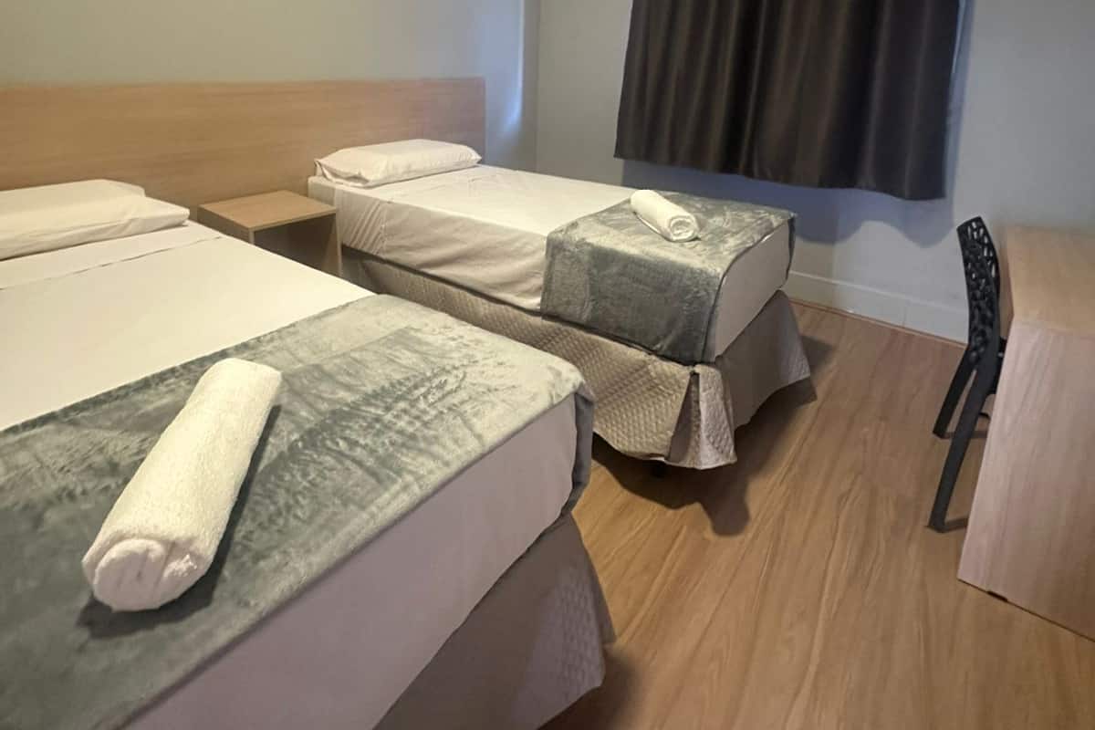 A hotel room with two beds, a wooden headboard, a small table, a chair, and a window with curtains.