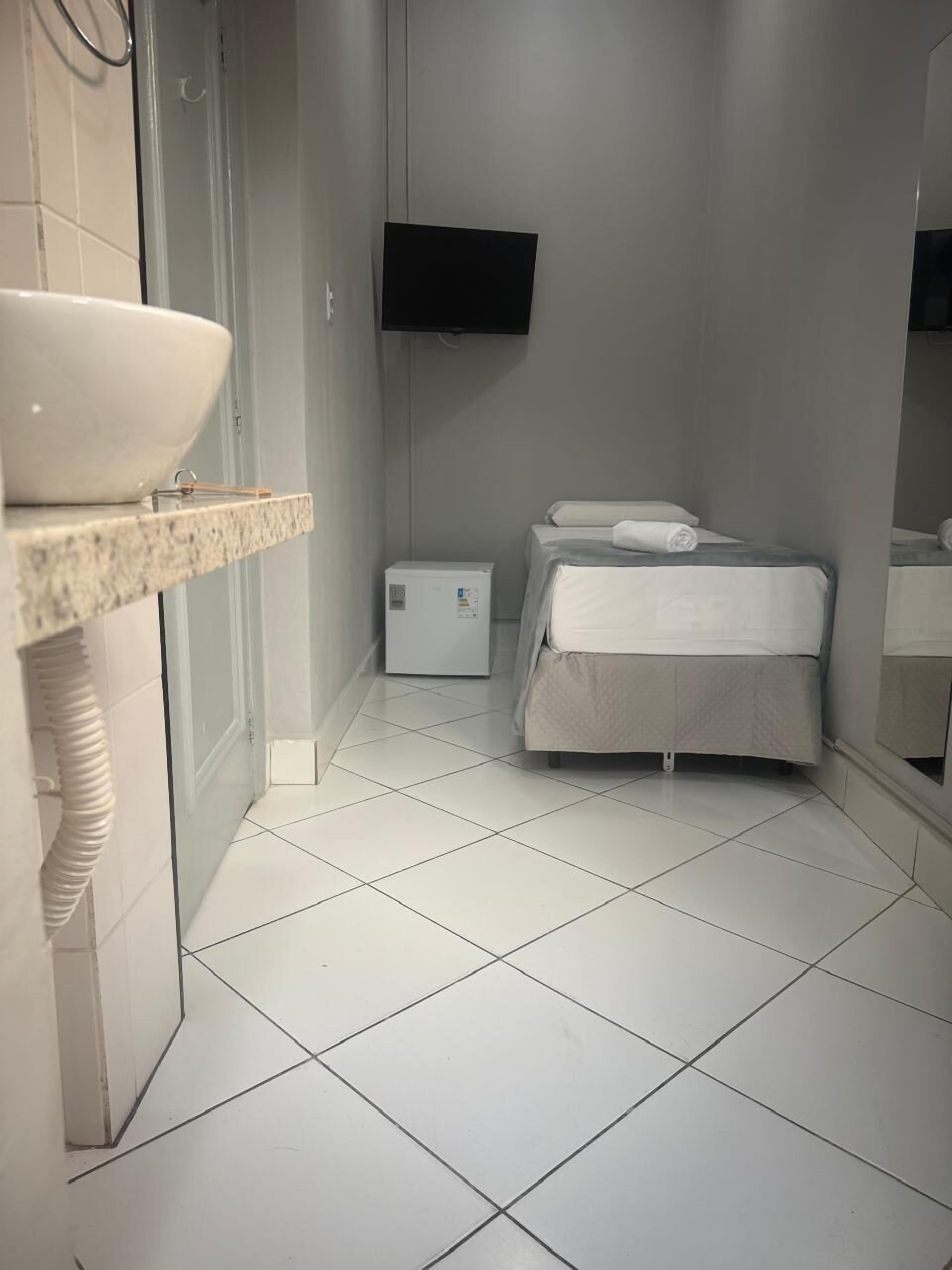 Un baño pequeño y moderno con un lavamanos blanco, una cama compacta y un televisor de pantalla plana montado en la pared.