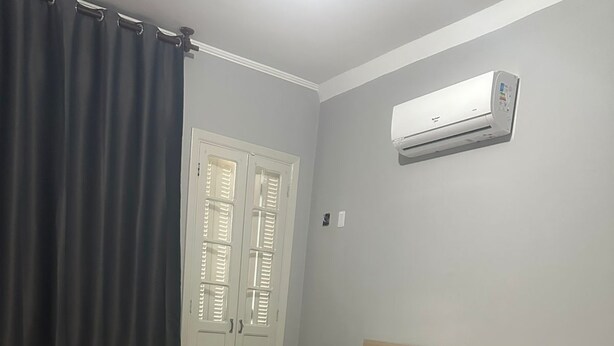 A bedroom with a bed, a nightstand, an air conditioning unit, a window with curtains, and a door with shutters. - Unity Hotel - Santos (Santos)